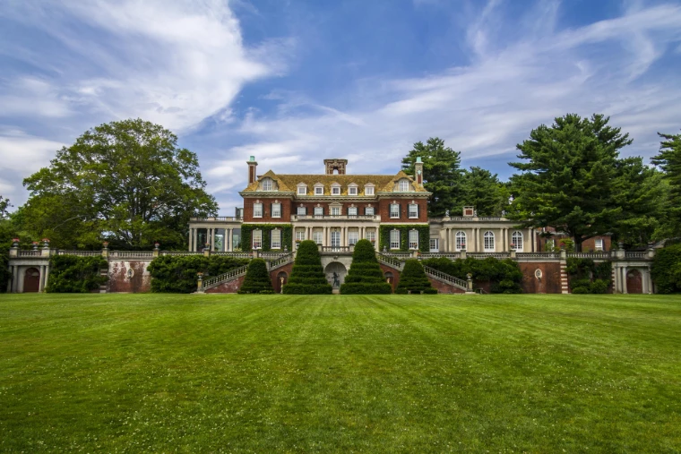 A red-brick Georgian-style mansion with white balustrades sits beyond a broad striped lawn and topiary under a blue sky with wispy clouds (2012).