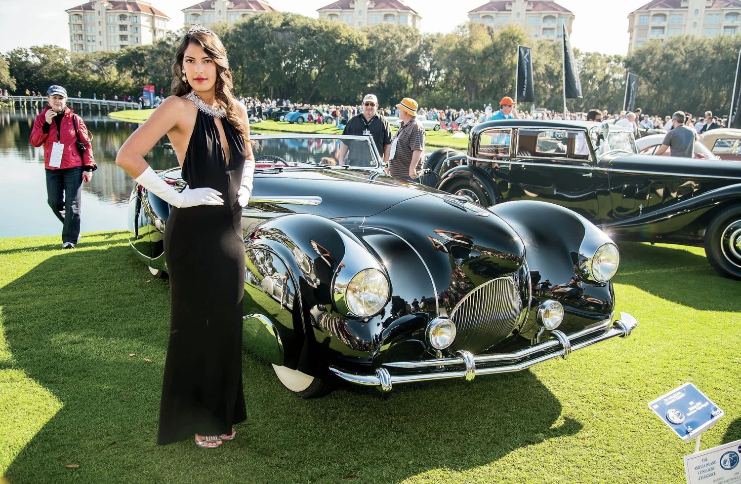 A guest in a black evening gown and white gloves beside a black prewar car on the show field at the Amelia Island Concours d’Elegance