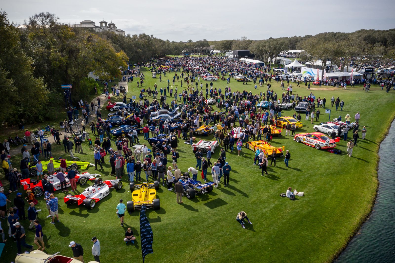 Racing cars ranked across the fairway with the crowd walking between them, seen from above at the Amelia Island Concours d’Elegance