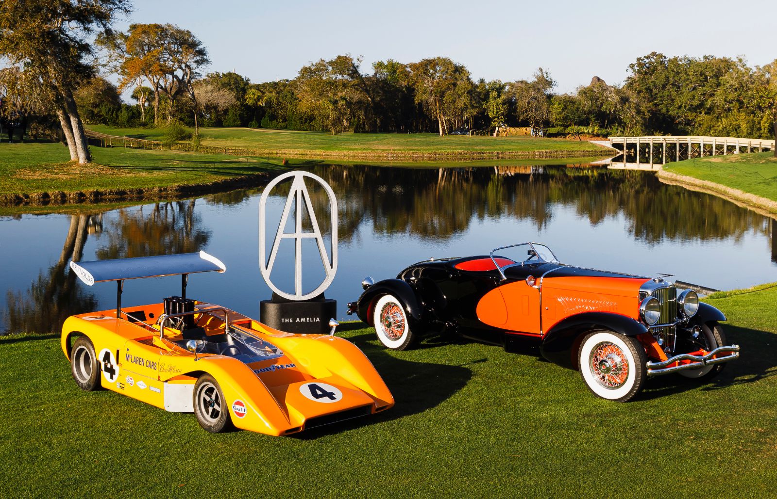An orange Can-Am McLaren and a prewar speedster flanking the Amelia trophy beside the lake at the Amelia Island Concours d’Elegance