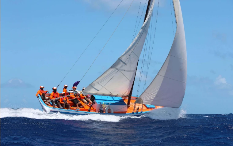 A traditional Caribbean workboat, crewed in orange, driving through the waves at the Antigua Classic Yacht Regatta, a mid-April engagement in the season calendar of The Mansa Court