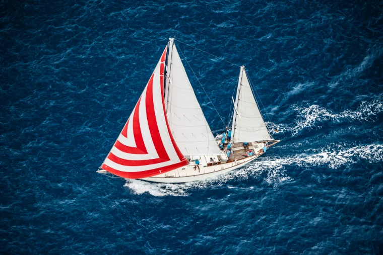 A classic ketch flying a red-and-white striped spinnaker, seen from above over deep blue water, at the Antigua Classic Yacht Regatta, a mid-April engagement in the season calendar of The Mansa Court