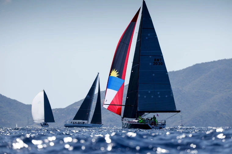 A racing yacht flying a spinnaker in the colours of the Antiguan flag, dark-sailed boats and the island hills beyond, at Antigua Sailing Week, a late-April engagement in the season calendar of The Mansa Court
