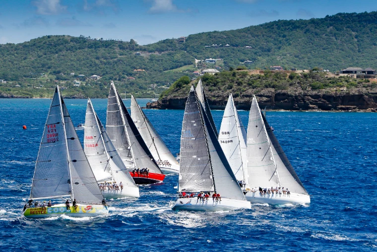 A fleet of racing yachts close-hauled together off the green hills of the Antigua coast at Antigua Sailing Week, a late-April engagement in the season calendar of The Mansa Court