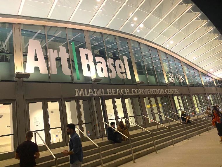 The Miami Beach Convention Center at dusk, “Art Basel” in large letters across its lit glass front above a long flight of steps, a few visitors outside (2021).
