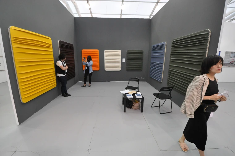 A gallery stand under a translucent ceiling hung with ridged wall works in yellow, orange, cream, grey and blue, a woman walking past in the foreground and two visitors looking on (2018).