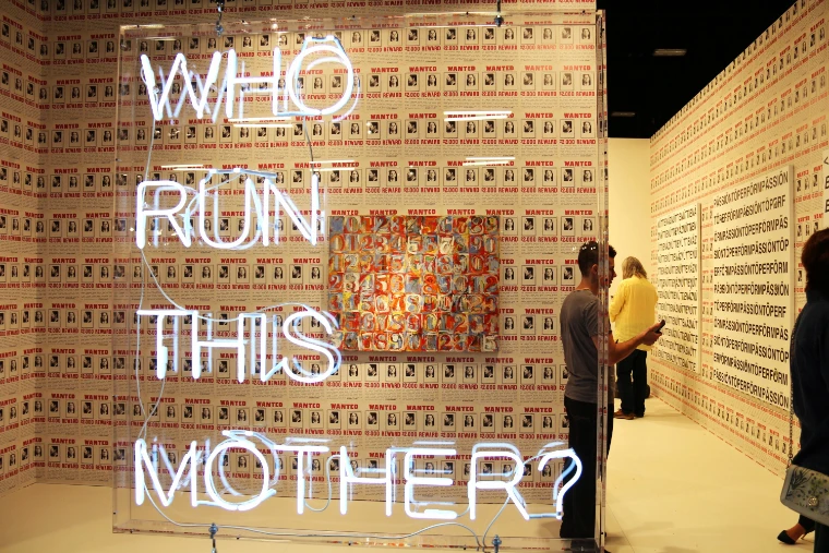 A white neon work reading “Who run this mother?” hangs before a booth papered in repeated posters, with visitors looking at the walls beyond (2015).