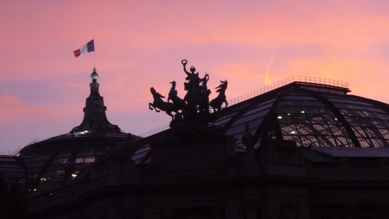 The bronze quadriga and glass roof of the Grand Palais in silhouette against a pink dusk sky, a French flag above (2007).