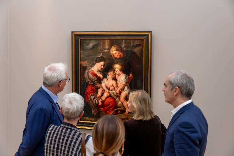 A small group of visitors gathers before an Old Master painting of a mother, children and an older woman, hung in a gilt frame on a gallery stand (2025).