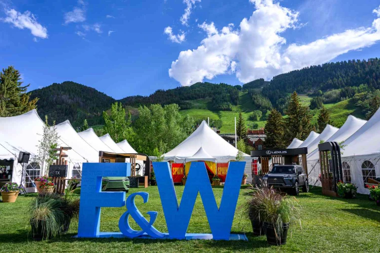 Tall blue letters on the festival lawn among white peaked tents, with the ski runs of Aspen Mountain rising behind, at the Aspen Food and Wine Classic, an engagement in the season calendar of The Mansa Court
