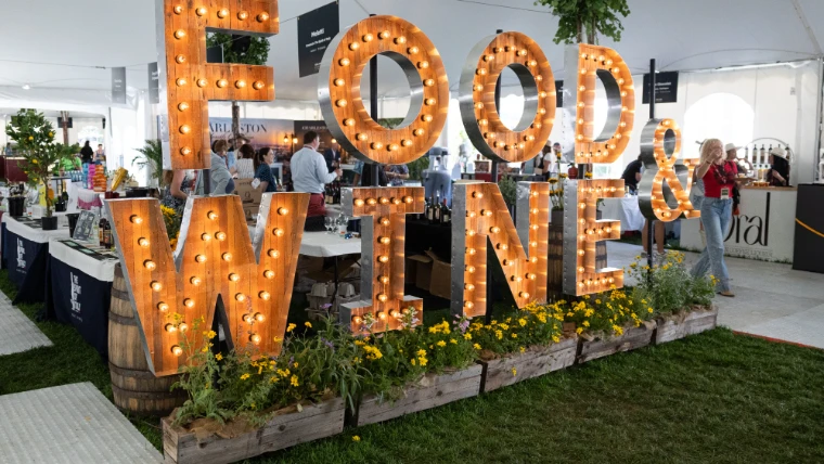 Marquee letters spelling Food & Wine above beds of wildflowers inside the Grand Tasting pavilion, wine and spirits stands beyond, at the Aspen Food and Wine Classic, a mid-June engagement in the season calendar of The Mansa Court