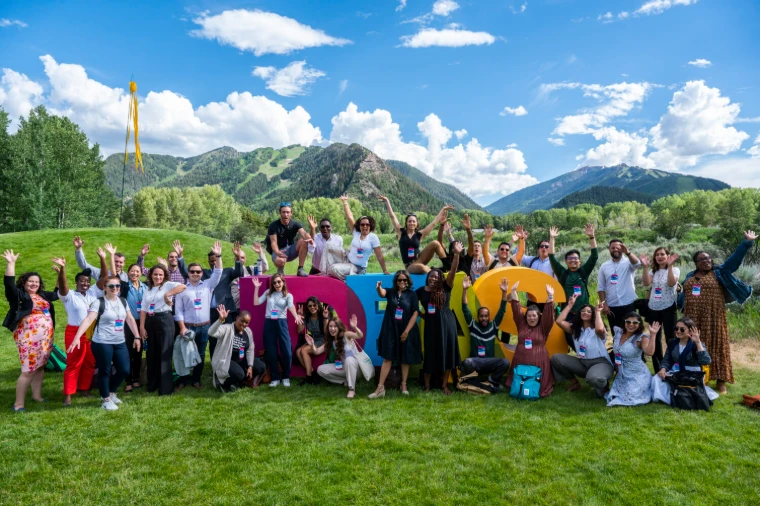 Festival guests gathered with arms raised around the giant Ideas letters on the meadow beneath the Rocky Mountains at the Aspen Ideas Festival, an engagement in the season calendar of The Mansa Court