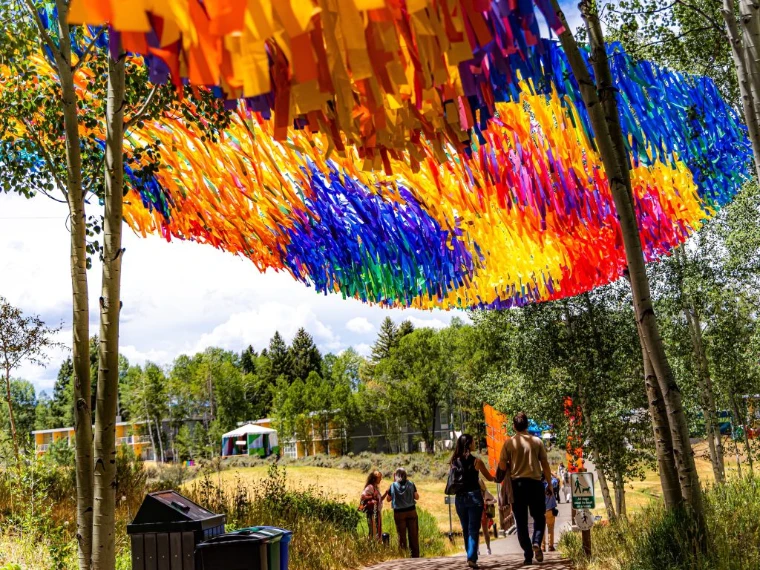 A canopy of coloured ribbons strung between aspen trees above a path through the Aspen Meadows campus at the Aspen Ideas Festival, the June festival in the season calendar of The Mansa Court