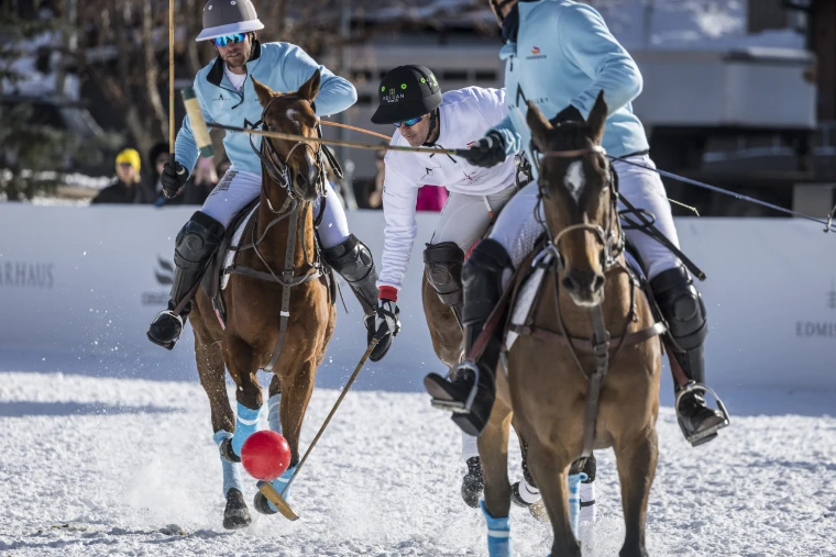 A red snow polo ball leaps into the air from a player’s mallet as riders in pale blue and white close in on the snow (2024).