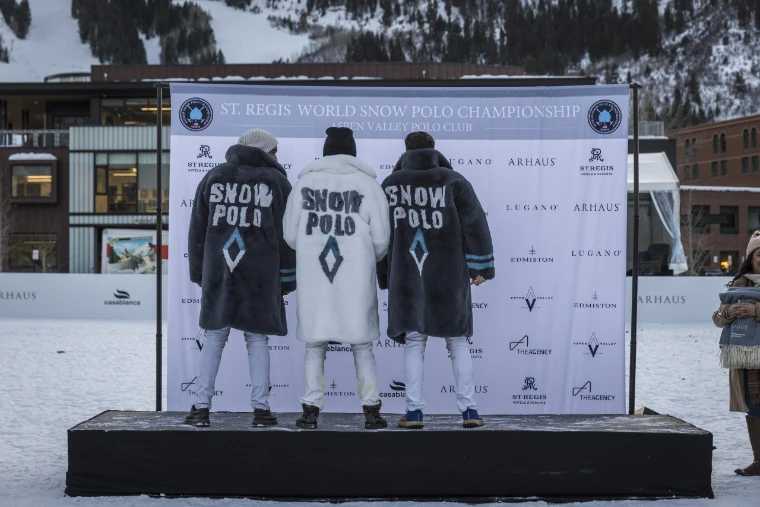 Three players stand on the prize-giving stage with their backs turned, wearing long fur coats lettered SNOW POLO, before the championship backdrop (2024).