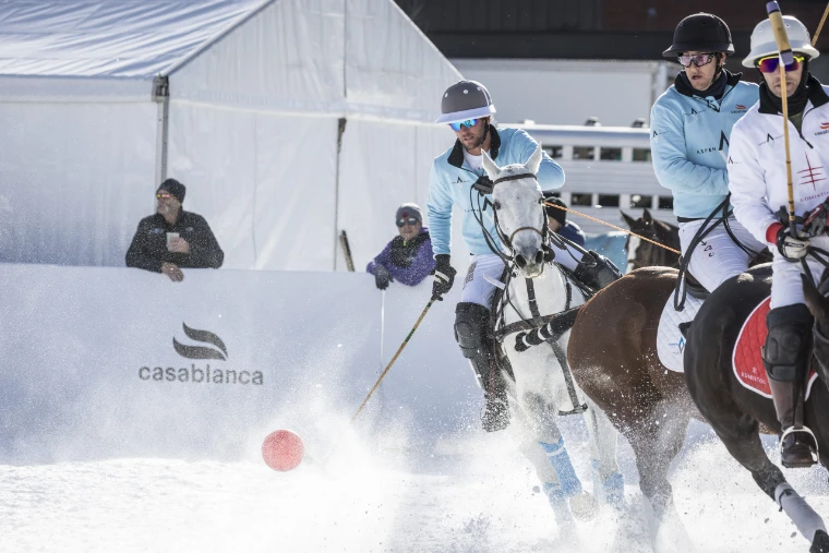 Riders in pale blue and white jerseys race a red ball along the boards in a spray of snow, spectators leaning over the barrier behind (2024).