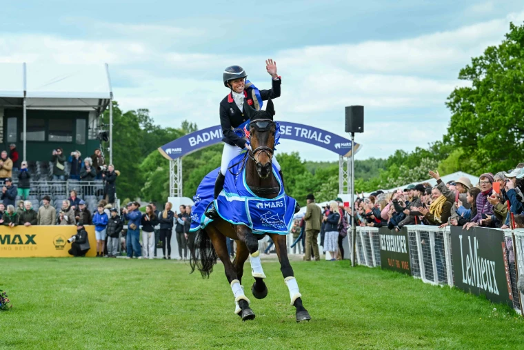 The winning rider waves to the crowd on her lap of honour, her horse in the winner’s rug beneath the Badminton arch, in 2026. Badminton Horse Trials, Gloucestershire, in the season calendar of The Mansa Court