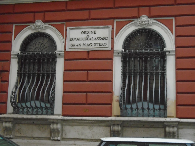 A stone plaque reading “Ordine dei SS. Maurizio e Lazzaro, Gran Magistero” between two barred, arched windows in a red façade (2017).