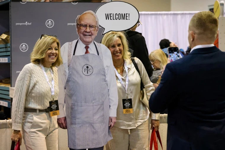 Two shareholders with guest passes pose beside a life-size cutout in an apron at a stand in the exhibition hall, in 2025. The Berkshire Hathaway Annual Meeting in Omaha, Nebraska, in the season calendar of The Mansa Court