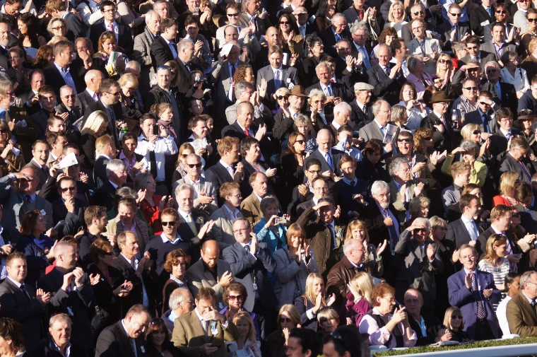 A packed crowd of racegoers in coats and suits applauds from the stands (2011).
