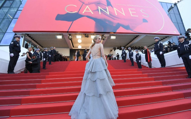 A guest in a tiered grey gown climbs the red-carpeted steps between the Palais guard of honour. The Cannes Film Festival at the Palais des Festivals, in the season calendar of The Mansa Court