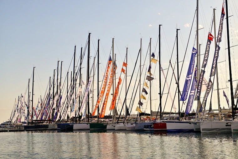 A long row of sailing-yacht masts hung with brand pennants and flags, their hulls moored along a pontoon in soft evening light (2026).