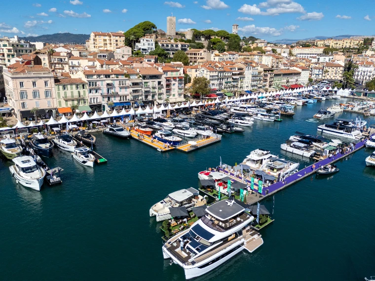 A raised view over the Vieux Port packed with moored motor yachts and catamarans on show, white tents along the quay, and the old town of Le Suquet with its tower on the hill behind (2026).