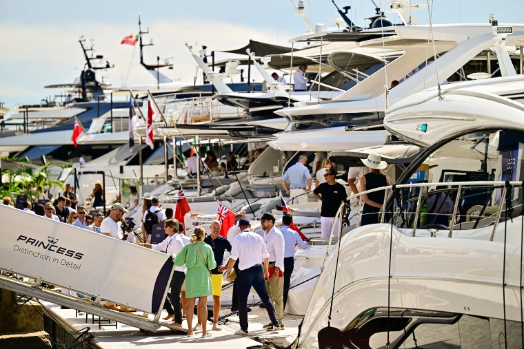 Visitors gather on the quay at the foot of a Princess yacht’s boarding passerelle, with a row of large white motor yachts flying red ensigns alongside (2026).