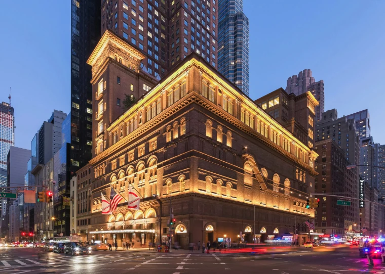 Carnegie Hall’s terracotta facade lit gold at dusk, flags over the entrance on Seventh Avenue. 57th Street and Seventh Avenue, New York, shown for Carnegie Hall Opening Night Gala in the season calendar of The Mansa Court