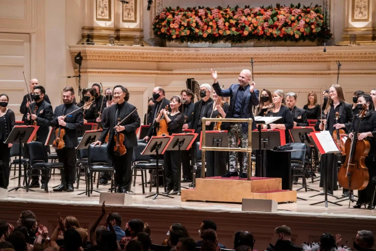 The orchestra stands to applause beneath a bank of autumn flowers on the Stern Auditorium stage at Carnegie Hall’s Opening Night Gala (2021). Carnegie Hall Opening Night Gala, Stern Auditorium, Carnegie Hall, New York, in the season calendar of The Mansa Court