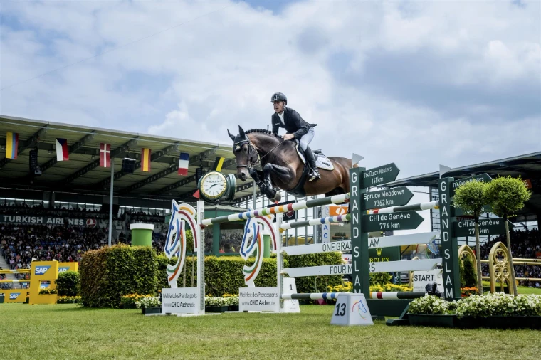 A bay horse jumping the Grand Slam fence beside the Rolex clock, signposts pointing to the other Majors, at CHIO Aachen, an engagement in the season calendar of The Mansa Court
