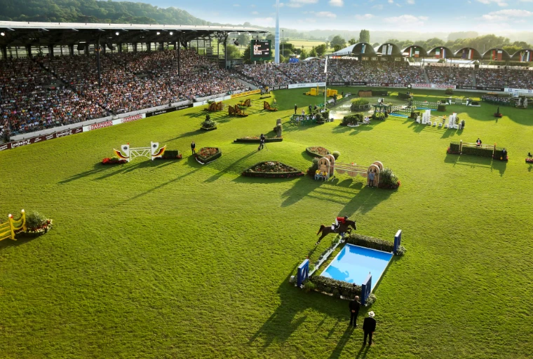 A rider clearing the water jump in the main stadium at the Soers in long evening light, the flag-topped stands full beyond, at CHIO Aachen, a July engagement in the season calendar of The Mansa Court
