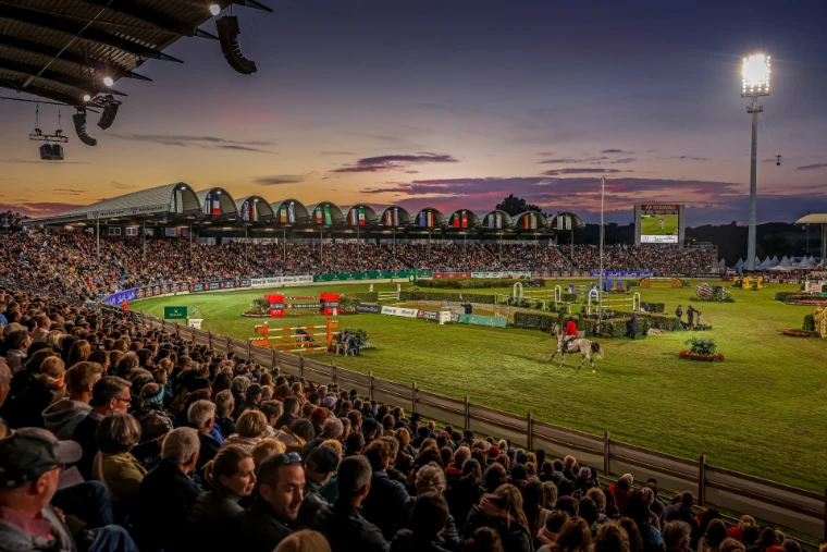 A packed stadium at dusk as a grey horse enters the floodlit arena beneath a violet sky at CHIO Aachen, the July show in the season calendar of The Mansa Court