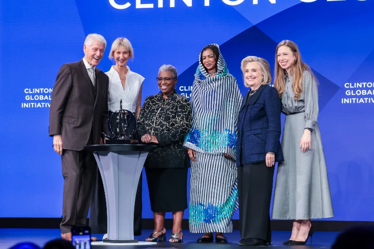 Six people pose smiling on a blue-lit stage beside an award on a pedestal, during the “Keep Going” mainstage session whose listed participants included Bill, Hillary and Chelsea Clinton (2026).