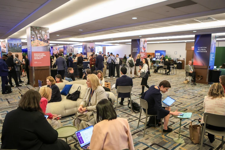 Delegates gather, talk and work on laptops among sofas and banner-wrapped pillars in a large hall of the meeting (2026).