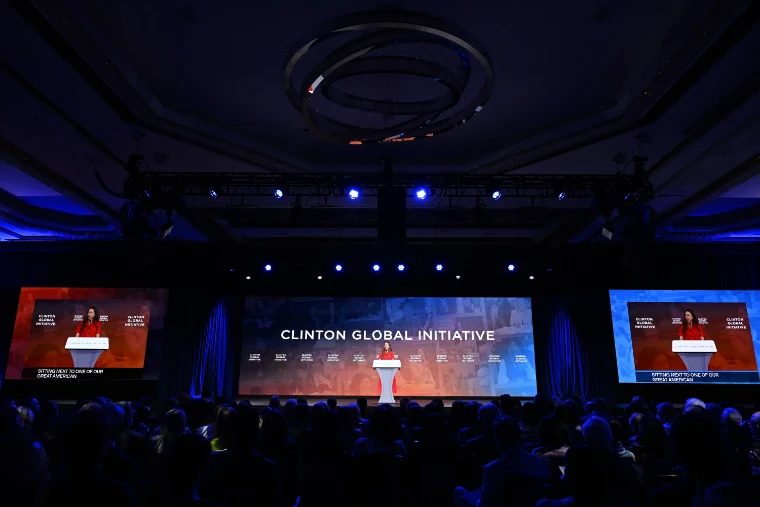 A darkened hotel ballroom full of seated delegates faces a wide stage where a speaker stands at a lectern beneath a “Clinton Global Initiative” screen, flanked by two large video screens and a ring-shaped chandelier overhead (2026).