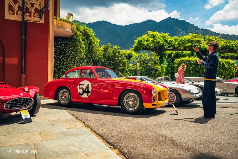 A red and yellow racing coupe numbered 54 beside the villa, a visitor photographing the line of cars, in 2025. The Concorso d’Eleganza Villa d’Este on Lake Como, in the season calendar of The Mansa Court