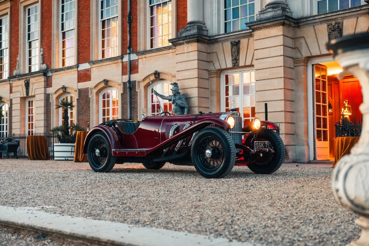 A dark red pre-war Mercedes-Benz SSK roadster, headlamps lit, on the gravel before the brick and stone front of the palace at dusk (2026).