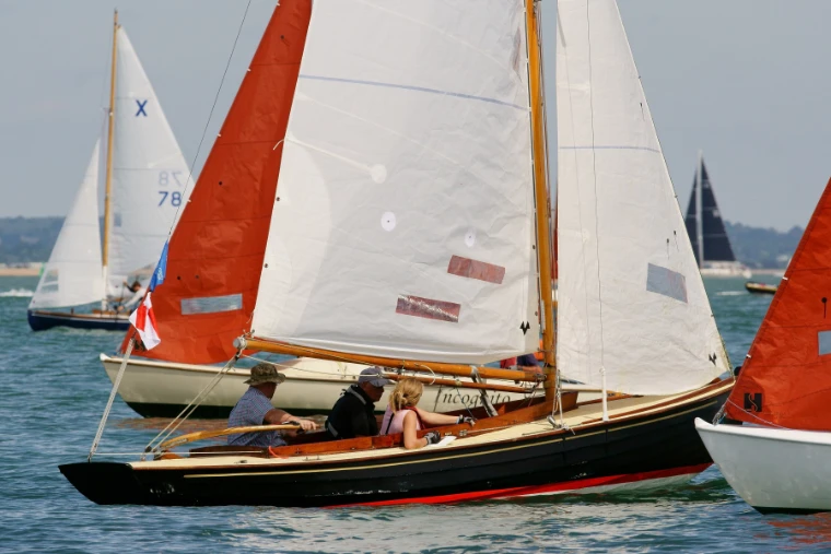 A crew sits in the cockpit of a varnished wooden keelboat with a white mainsail, between boats with orange sails (2018).