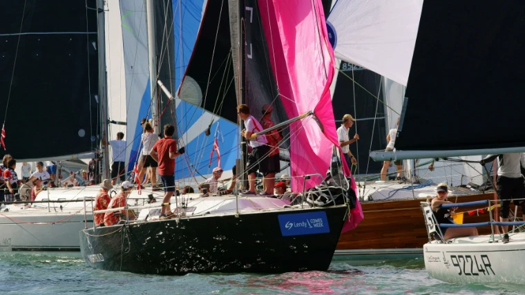 Crews stand on the decks of closely packed racing yachts under black, pink and white spinnakers (2018).