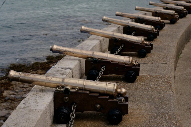 A row of brass starting cannons on wooden carriages lines a curved stone sea wall above the water (2019).