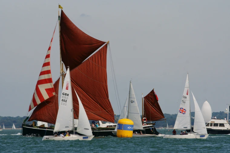 A Thames sailing barge with rust-red sails and a striped topsail sails among smaller white-sailed and red-sailed racing boats, with a yellow race mark in the water (2018).