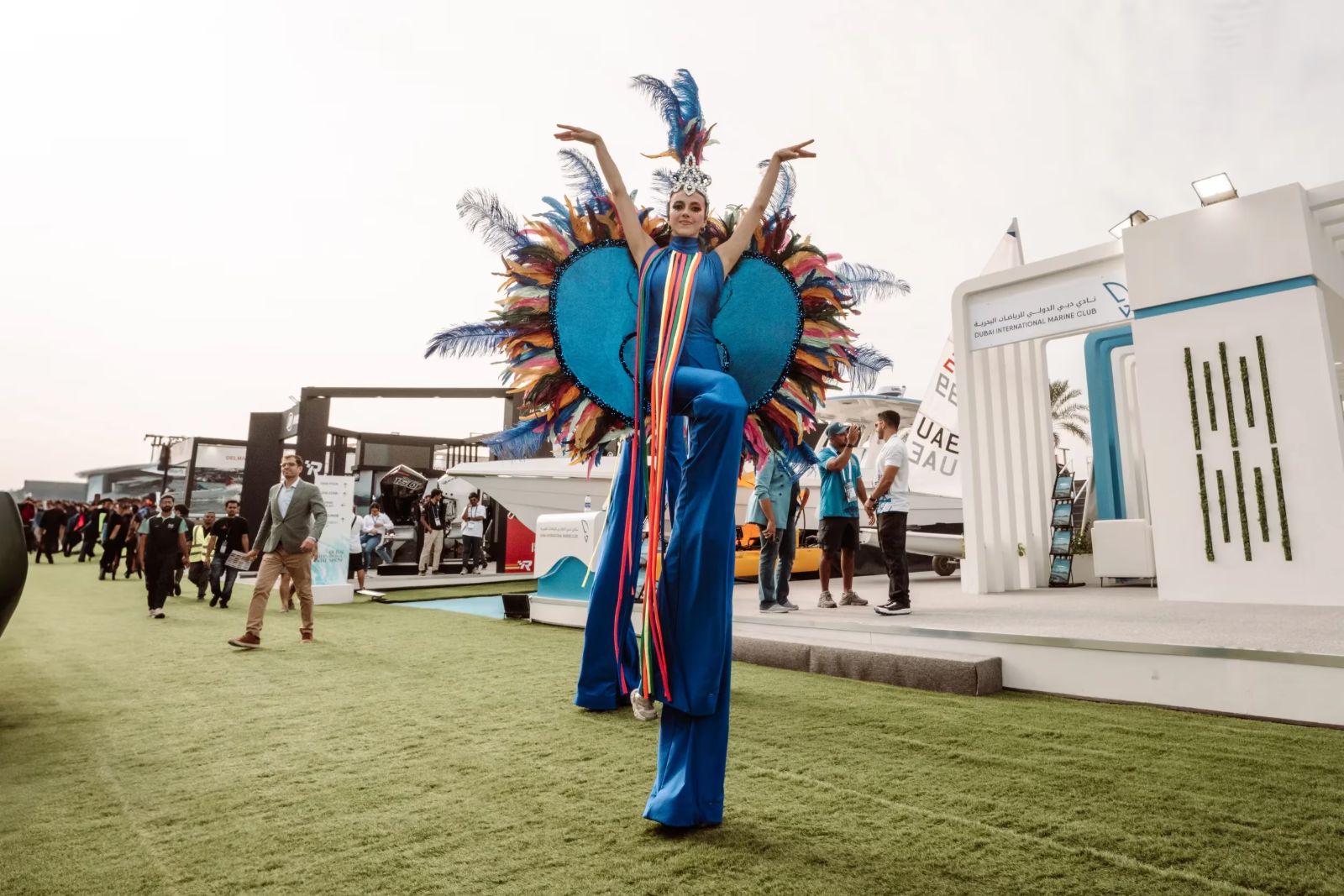 A stilt performer in feathered costume crossing the lawn between the stands at the Dubai International Boat Show