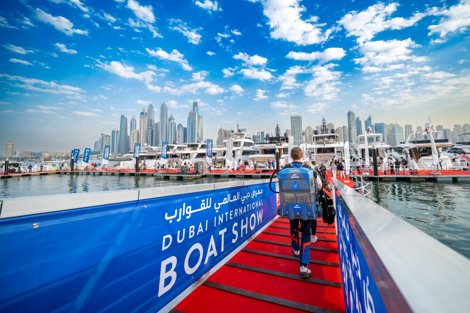 Visitors crossing onto the red-carpet pontoons past the show branding with yachts and the Dubai Marina skyline beyond at the Dubai International Boat Show