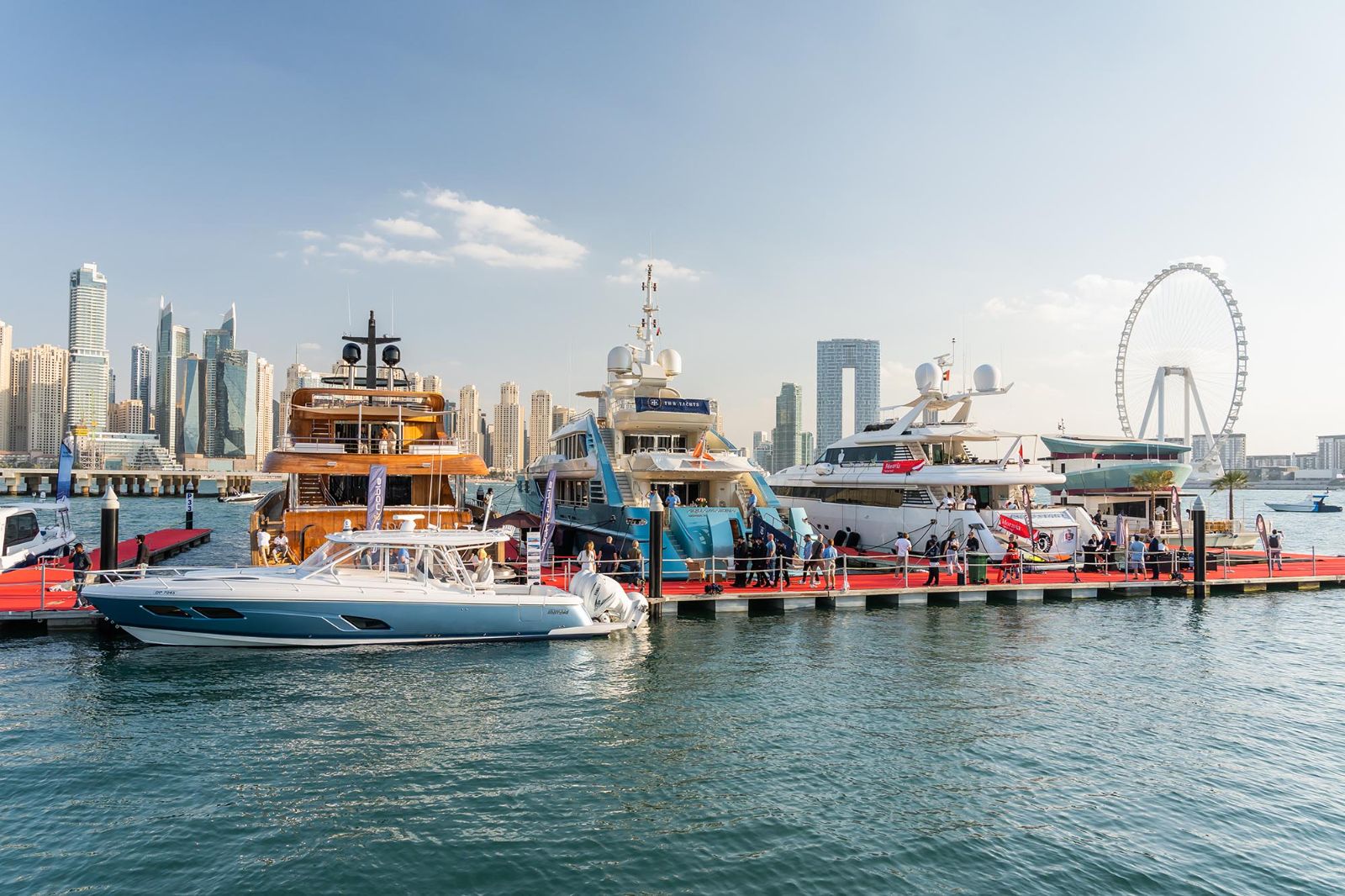 Superyachts and a classic wooden motor yacht berthed along the red pontoons with Ain Dubai beyond at the Dubai International Boat Show