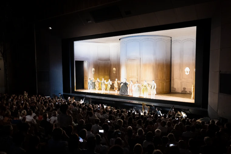 A full audience sits in the dark facing a brightly lit stage where performers stand before a pale curved set (2025).