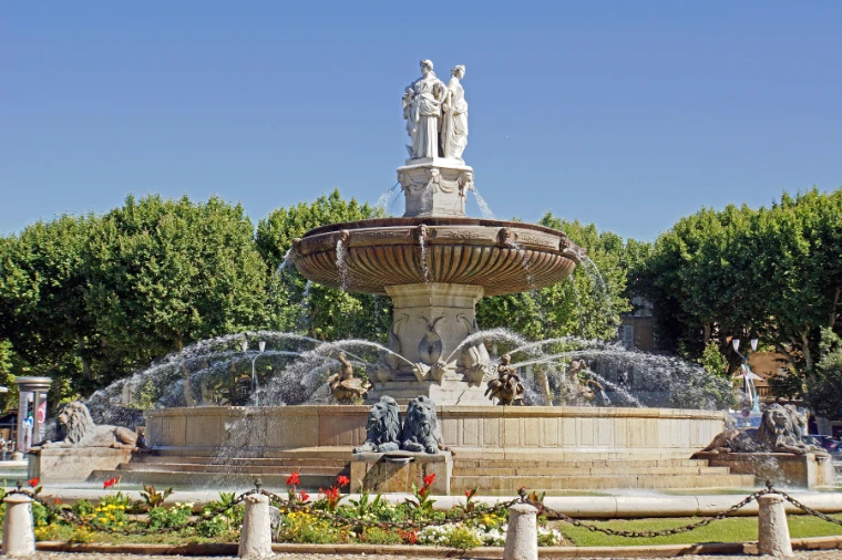 The tiered Fontaine de la Rotonde sprays water beneath three marble statues, ringed by flowerbeds and green summer trees under a blue sky (2014).