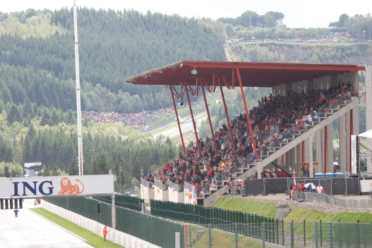 A packed grandstand with a red steel roof overlooks the circuit, with forested Ardennes hills and more spectators on the slopes beyond (2008).