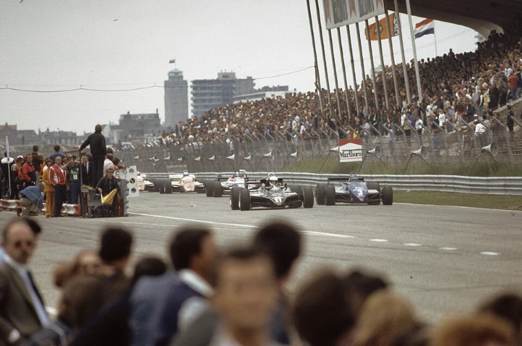Racing cars pull away at the start before a packed grandstand hung with flags, spectators’ heads in the foreground (1982).