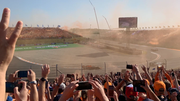 Fans with raised hands and phones fill the foreground as orange smoke drifts over the banked corner and packed grandstands of the circuit (2021).